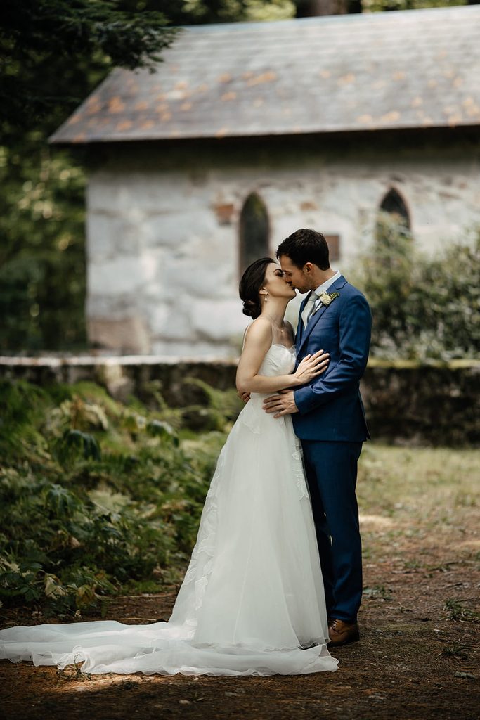Caroline and Arthur kissing in the woods beside the stone chapel