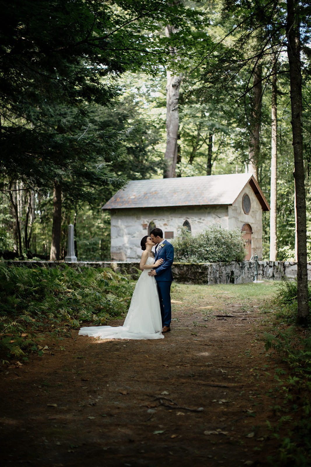 Caroline and Arthur kissing on a wooded path beside a stone chapel