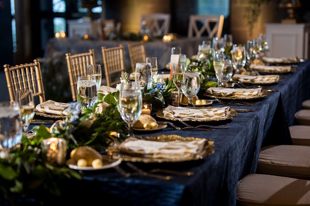 Navy reception table with gold place settings, greenery, and candlelight